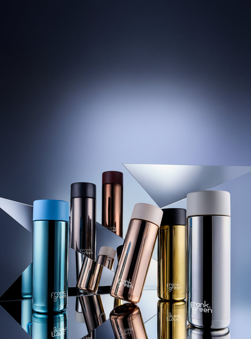 A group of frank green travel mugs in various metallic colors including blue, rose gold, black, gold, and silver, arranged on a reflective surface with geometric background props.