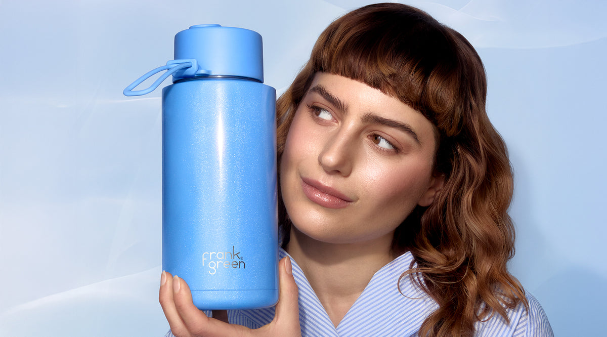 A woman with wavy brown hair and bangs holds a tall, light blue water bottle with a matching blue cap and carrying loop. The bottle has the brand name 'franks fighen' printed near the bottom.