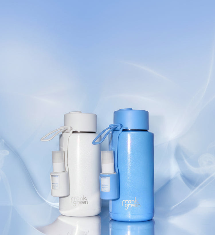 Two insulated water bottles from frank green on a reflective light blue surface. The left bottle is matte white with a white cap and a small white bottle accessory attached.